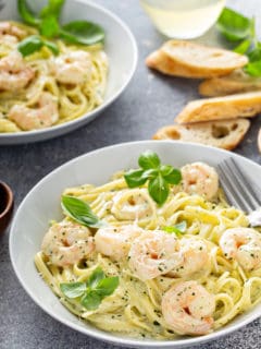 Plated bowl of creamy pesto pasta with shrimp with another bowl and slices of baguette in the background