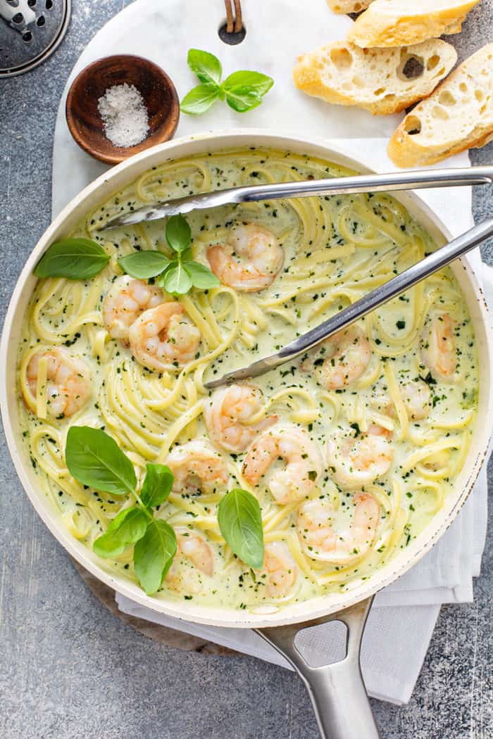 Metal tongs tossing together creamy pesto pasta sauce with pasta and shrimp in a saucepan