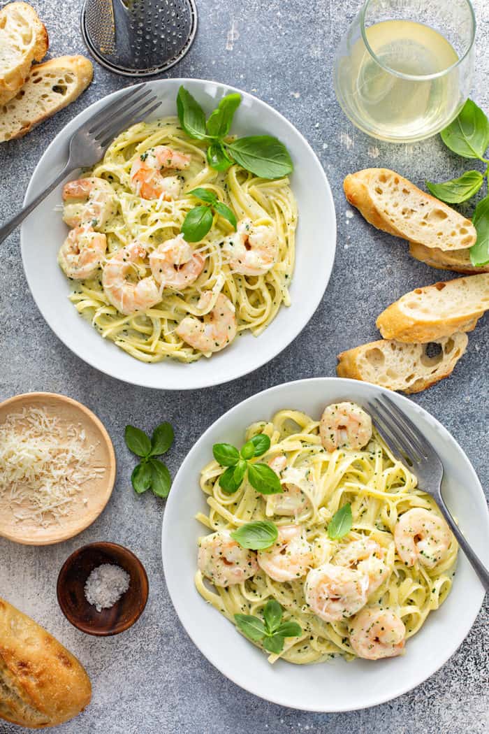 Overhead view of two white bowls filled with creamy pesto pasta with shrimp, set alongside slices of baguette and bowls of parmesan cheese