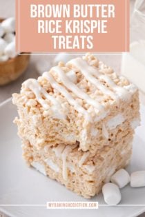 Two brown butter rice krispie treats stacked on a white plate. The top treat has a bite taken out of the corner. Text overlay includes recipe name.