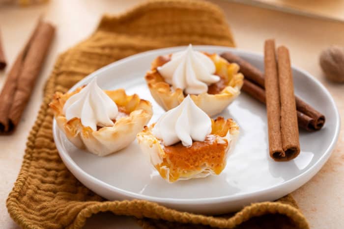 White plate holding mini pumpkin pies next to cinnamon sticks. The pie in the front is cut in half to show the filling.
