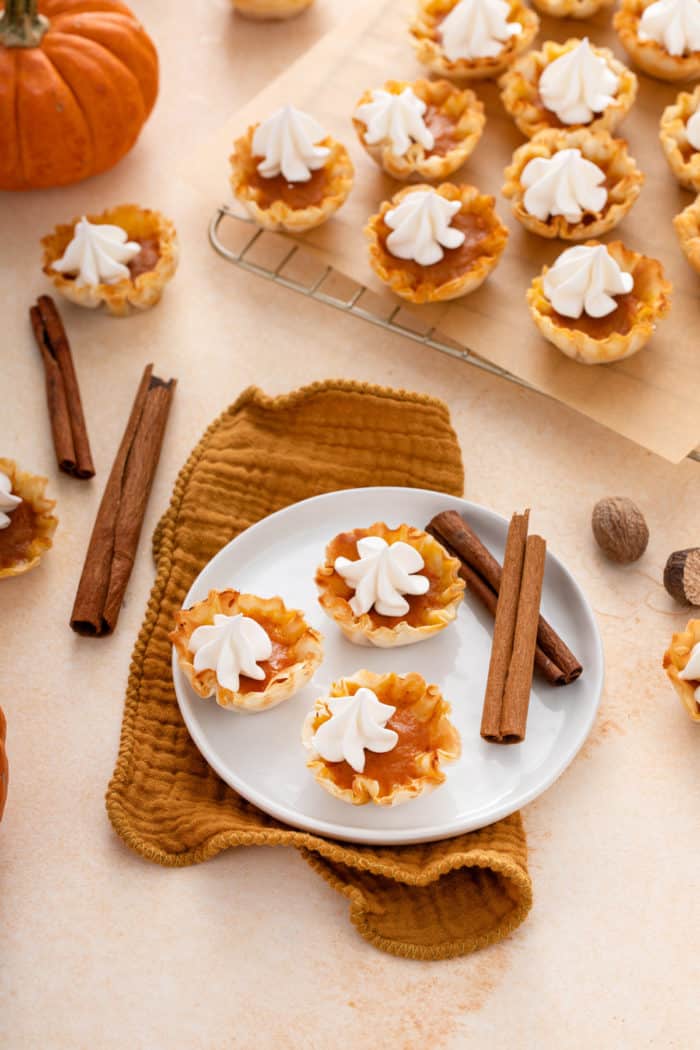 White plated holding three mini pumpkin pies on a brown napkin in front of a wire rack holding more mini pumpkin pies.