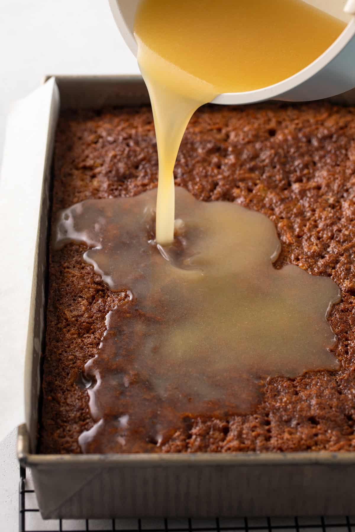 buttermilk syrup being poured over a baked carrot cake in a 9x13-inch pan.