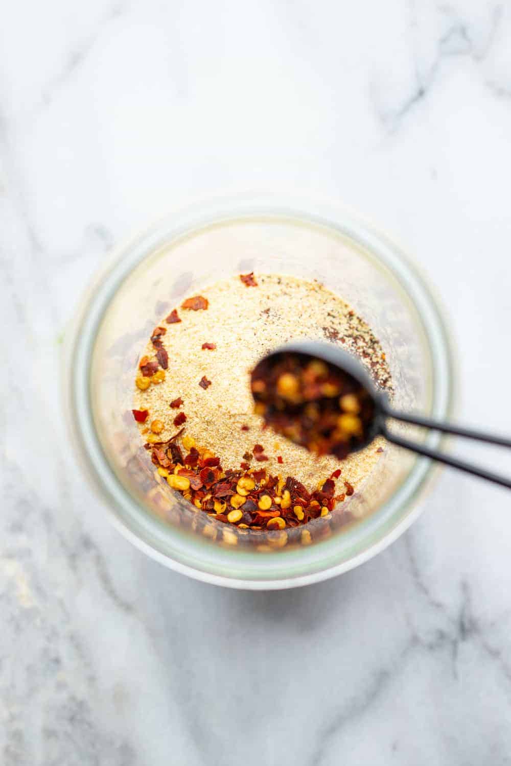 Measuring spoon adding crushed red pepper to a jar for taco seasoning