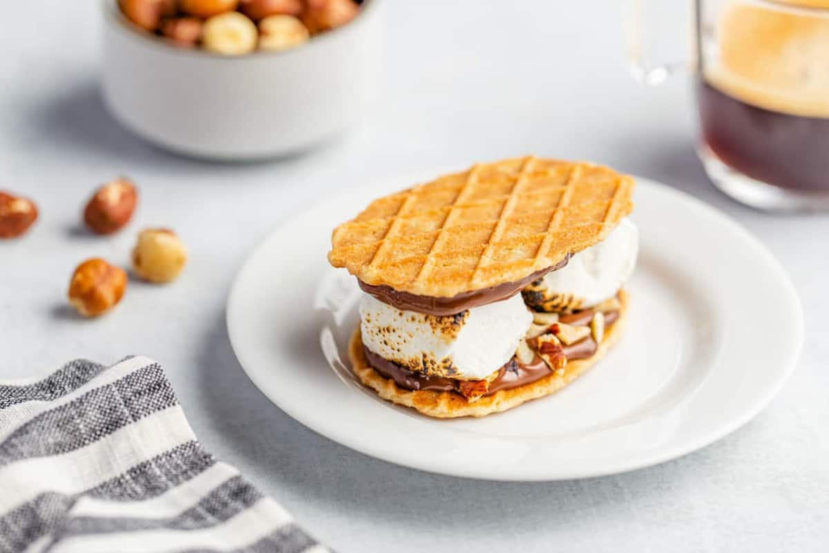 Plated s'more made with waffle cookies, chocolate hazelnut spread and 2 large roasted marshmallows with bowl of toasted hazelnuts in the background