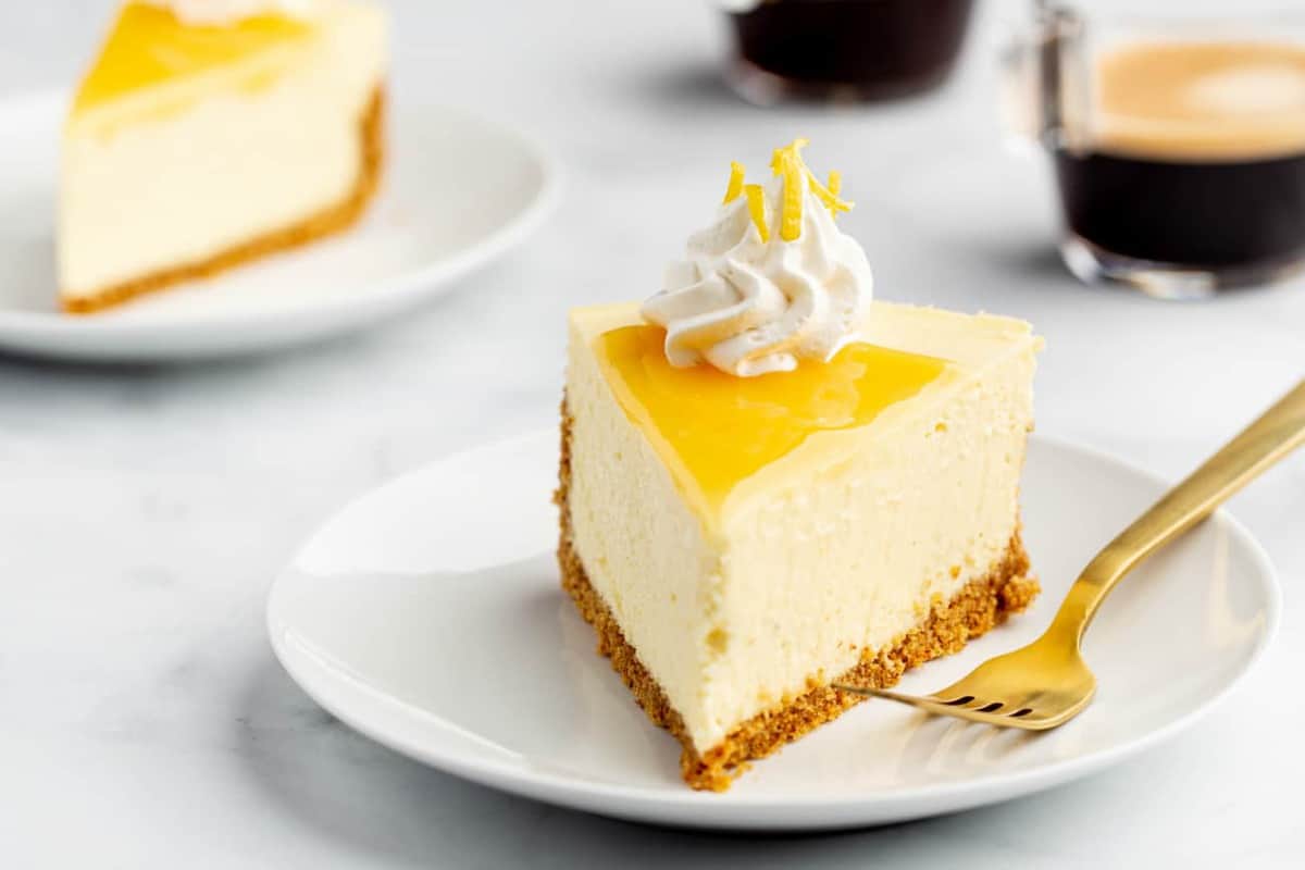 Plated slice of lemon cheesecake next to a fork with cup of espresso and another slice of cheesecake in the background