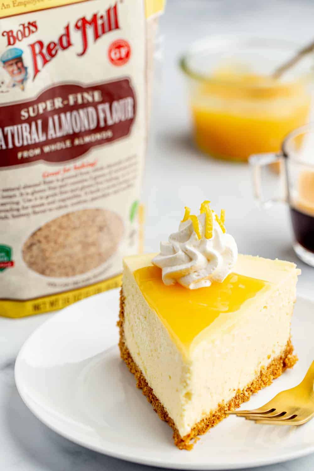 Plated lemon cheesecake next to bag of finely ground almond meal with bowl of lemon curd in the background