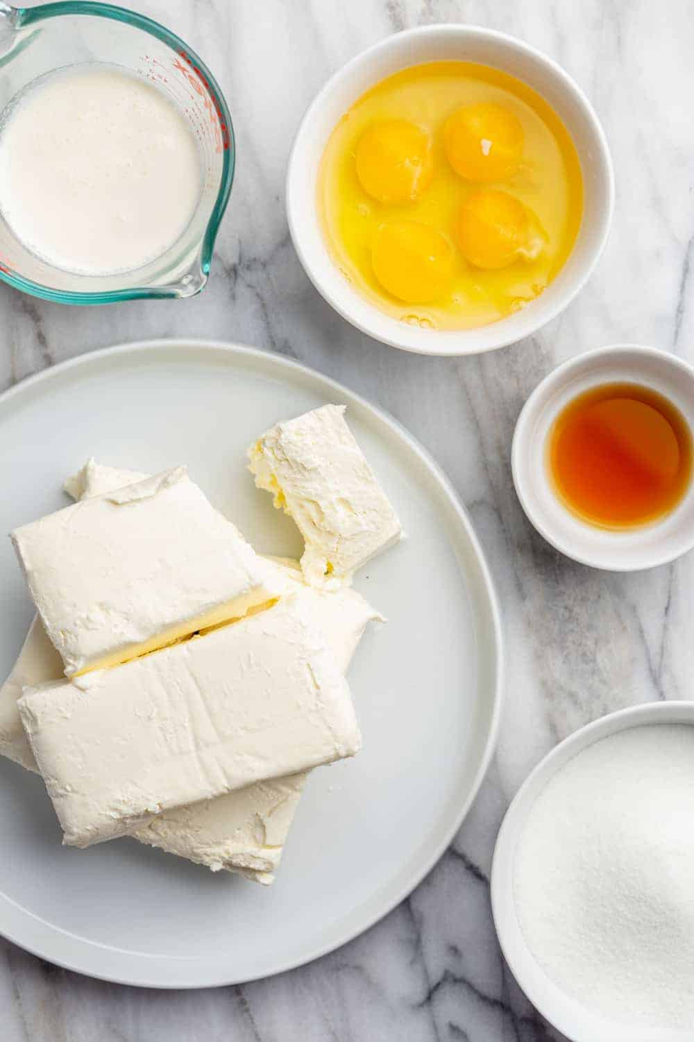 Blocks of cream cheese on a white plate, surrounded by cheesecake ingredients