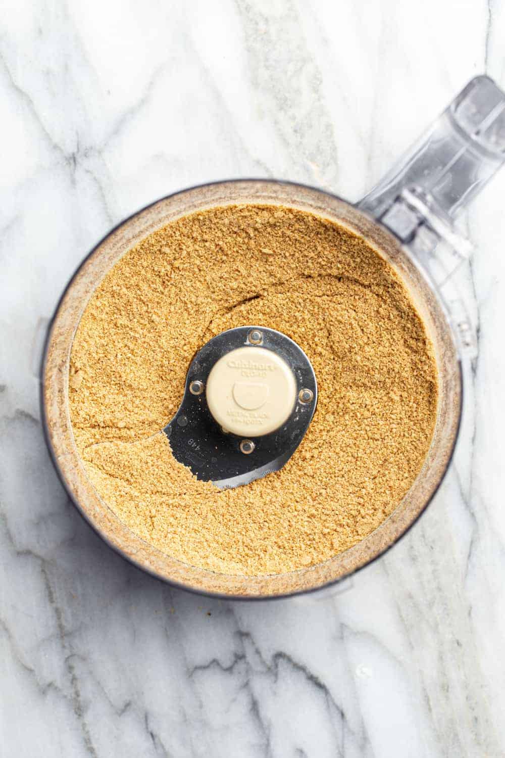 Graham cracker crumbs in the bowl of a food processor on a marble surface