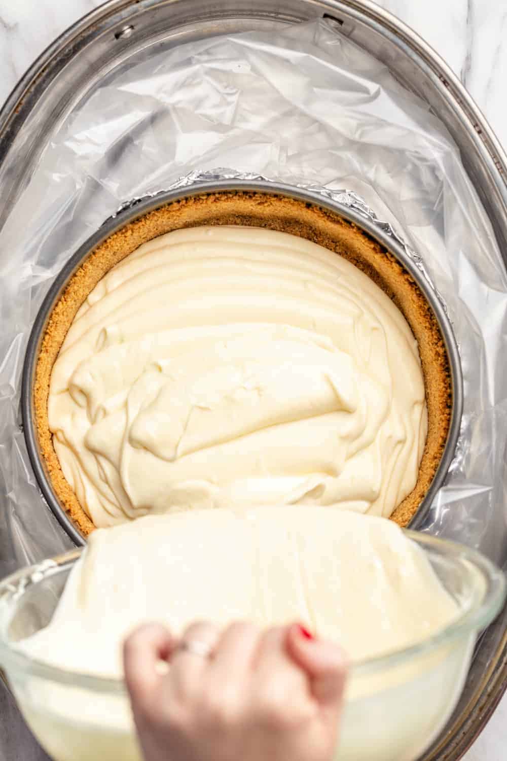 Pouring cheesecake filling into a graham cracker crust before adding a water bath