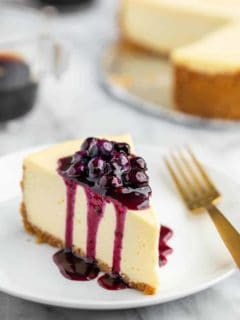 Slice of vanilla cheesecake topped with blueberry sauce, with the rest of the cheesecake in the background
