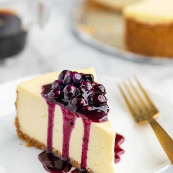 Slice of vanilla cheesecake topped with blueberry sauce, with the rest of the cheesecake in the background