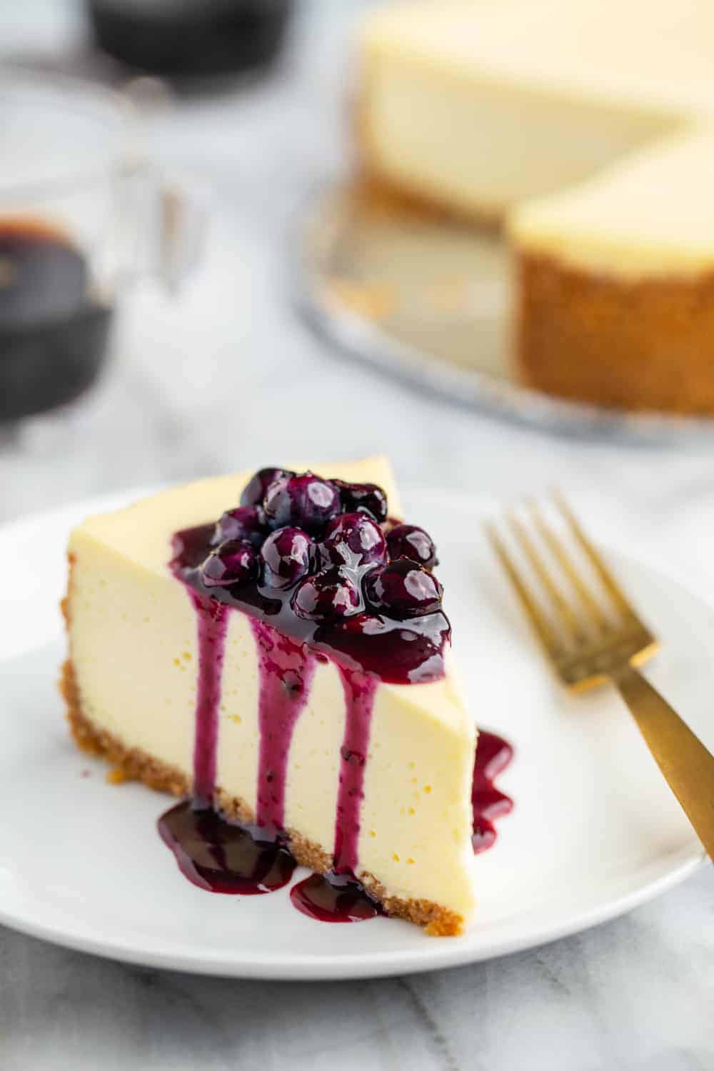 Slice of vanilla cheesecake topped with blueberry sauce, with the rest of the cheesecake in the background