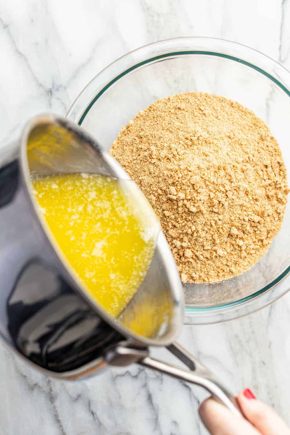 Pouring melted butter from a saucepan into a bowl of graham cracker crumbs