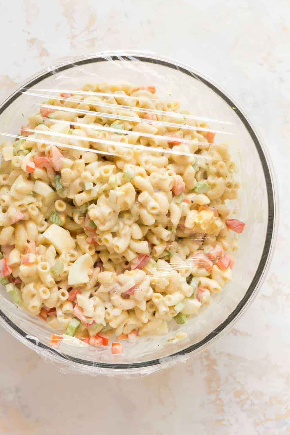 Macaroni salad in a glass bowl, covered with plastic wrap