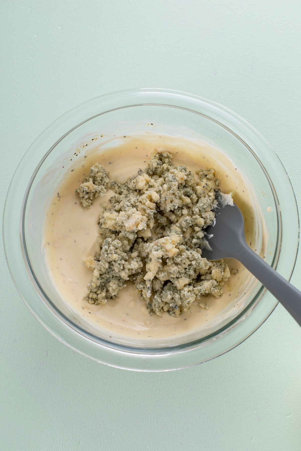 Blue cheese crumbles about to be stirred into a salad dressing base in a glass bowl.