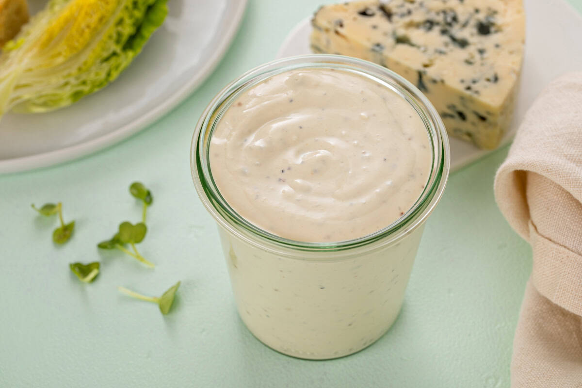 Top-down view of a glass jar filled with blue cheese dressing set next to a salad and a chunk of blue cheese on a countertop.