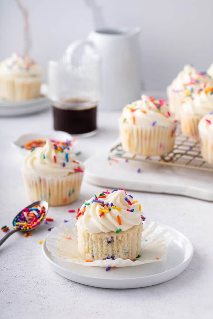 Unwrapped funfetti cupcake on a white plate with more cupcakes in the background.