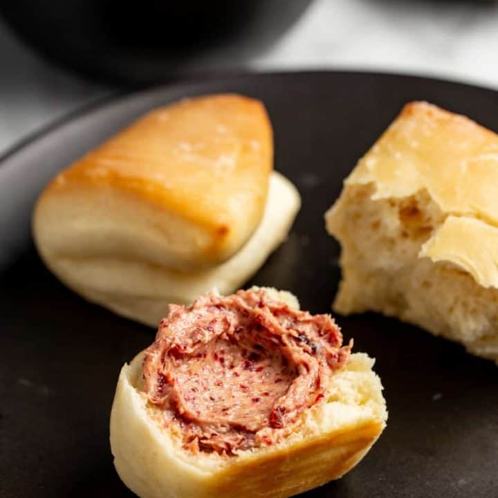 Parker house rolls on a plate, with one roll cut in half and spread with cranberry butter