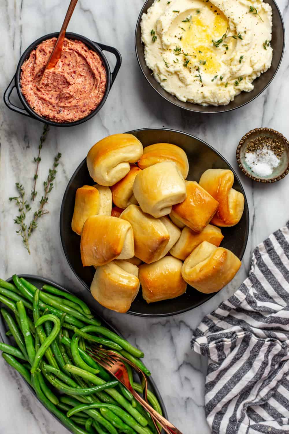 Thanksgiving dinner sides on a marble surface: a bowl of green beans, a bowl of mashed potatoes, and a small bowl of cranberry butter surrounding parker house rolls