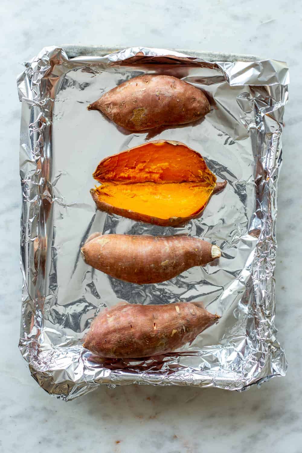 Baked sweet potatoes on a foil-lined sheet tray, with one of the sweet potatoes split open