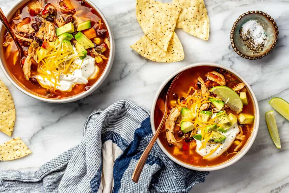 Two bowls of chicken tortilla soup alongside tortilla chips on a marble surface