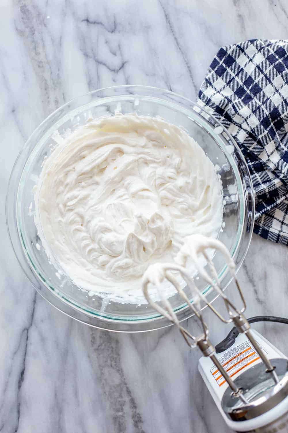 Glass mixing bowl of freshly whipped cream next to a hand mixer