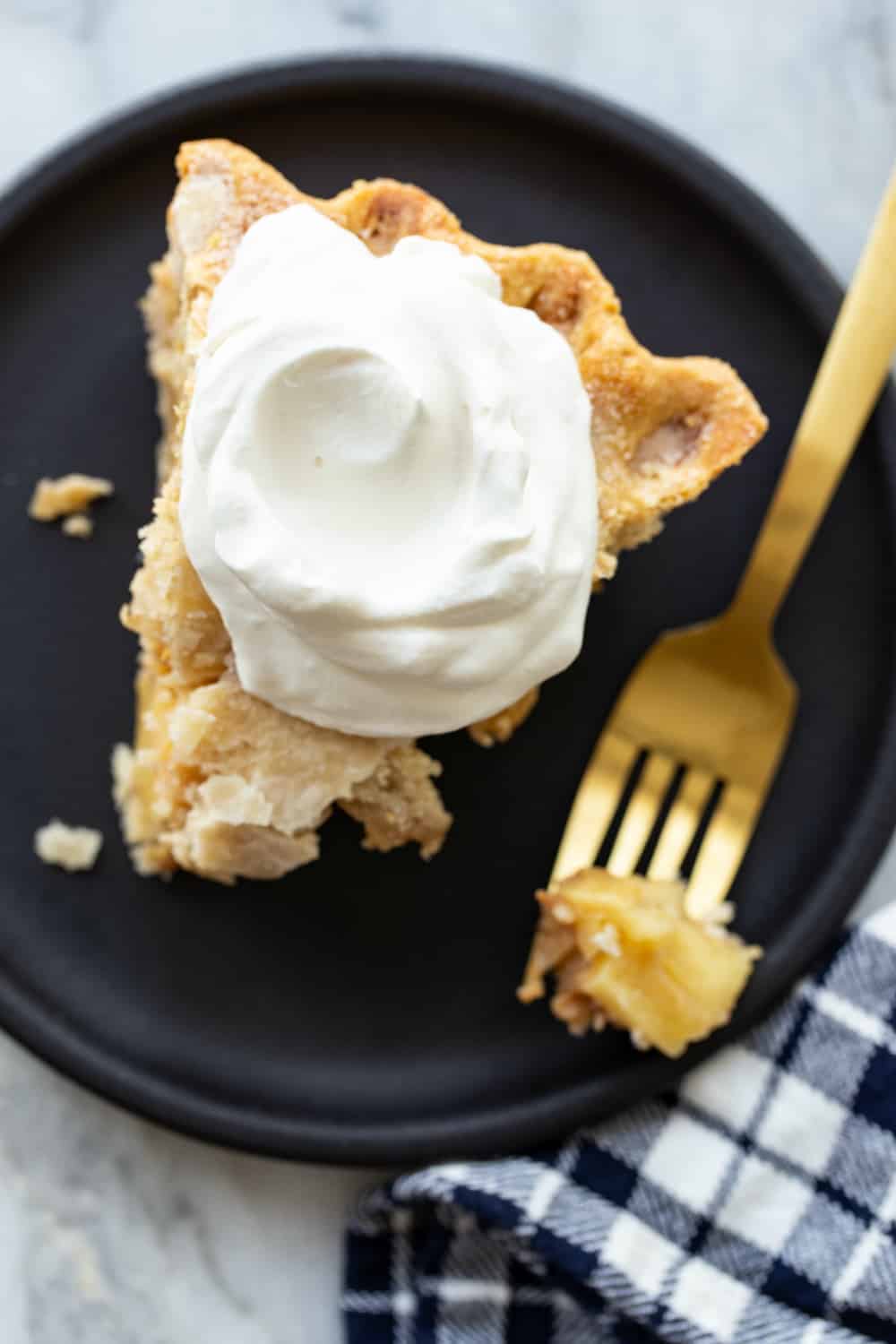 Slice of apple pie on a black plate, topped with softly whipped cream