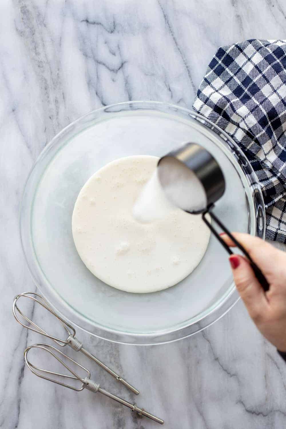 Hand pouring sugar into a glass mixing bowl of heavy cream to make whipped cream