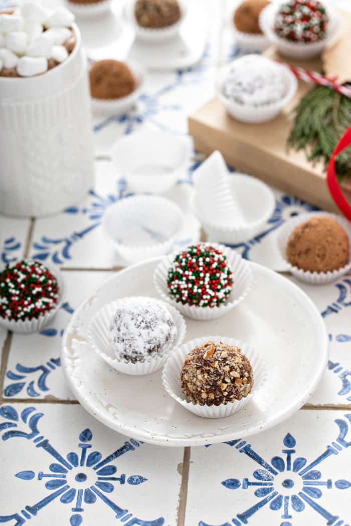 Three rum balls arranged on a white plate with more rum balls and a mug of hot cocoa in the background