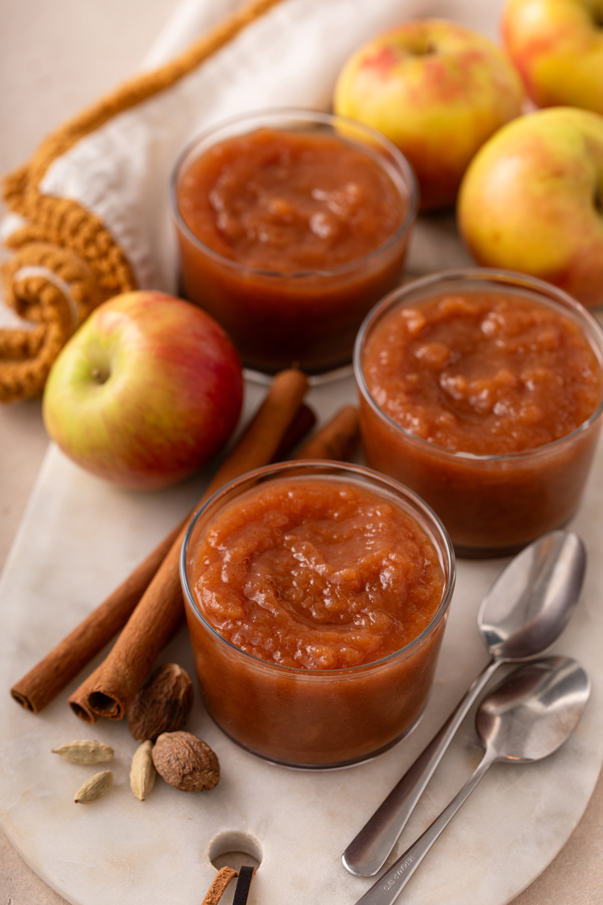 Three small bowls of crockpot applesauce next to whole apples and cinnamon sticks.
