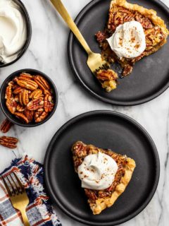 Slices of pecan pie on black plates arranged next to a bowl of pecans and a bowl of whipped cream