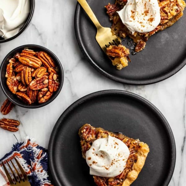 Slices of pecan pie on black plates arranged next to a bowl of pecans and a bowl of whipped cream