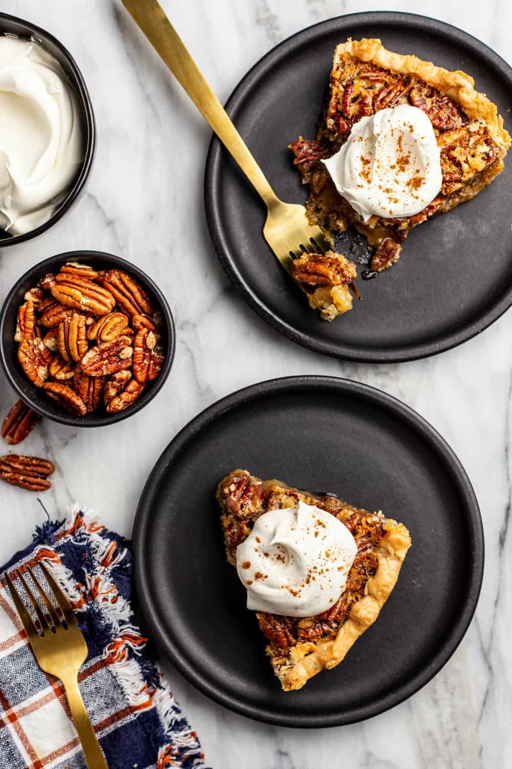 Slices of pecan pie on black plates arranged next to a bowl of pecans and a bowl of whipped cream