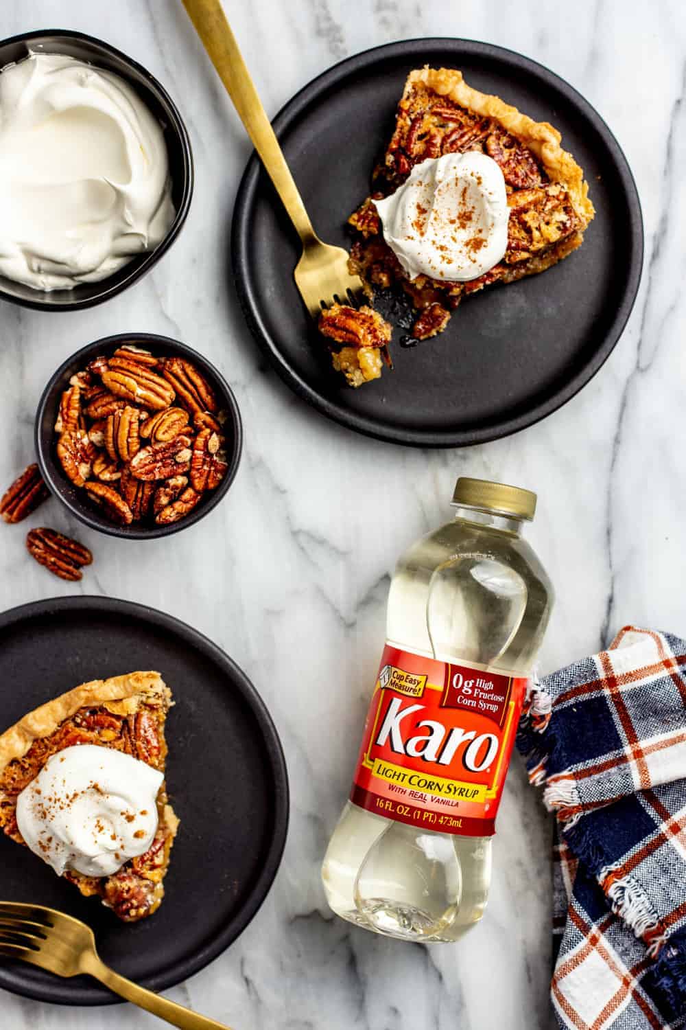 Overhead view of slices of pecan pie on black plates, arranged next to a bowl of pecans, a bowl of whipped cream and a bottle of corn syrup