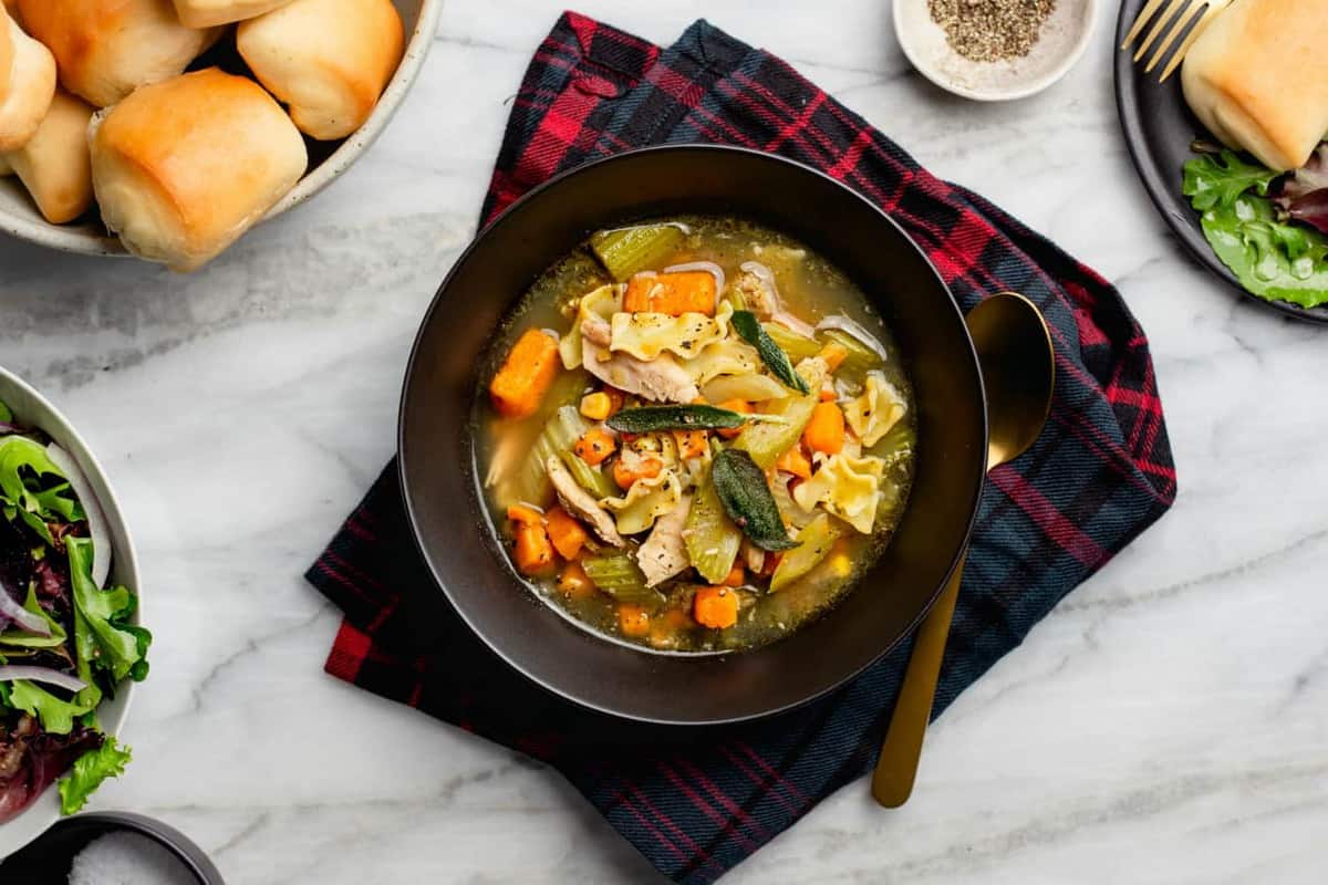 Turkey noodle soup in a black bowl on top of a plaid napkin on a marble surface