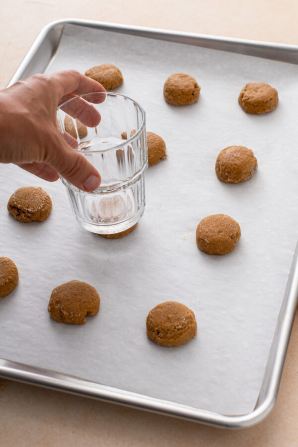 Person using the bottom of a drinking glass to press down balls of pumpkin molasses cookie dough on a parchment-lined baking sheet.