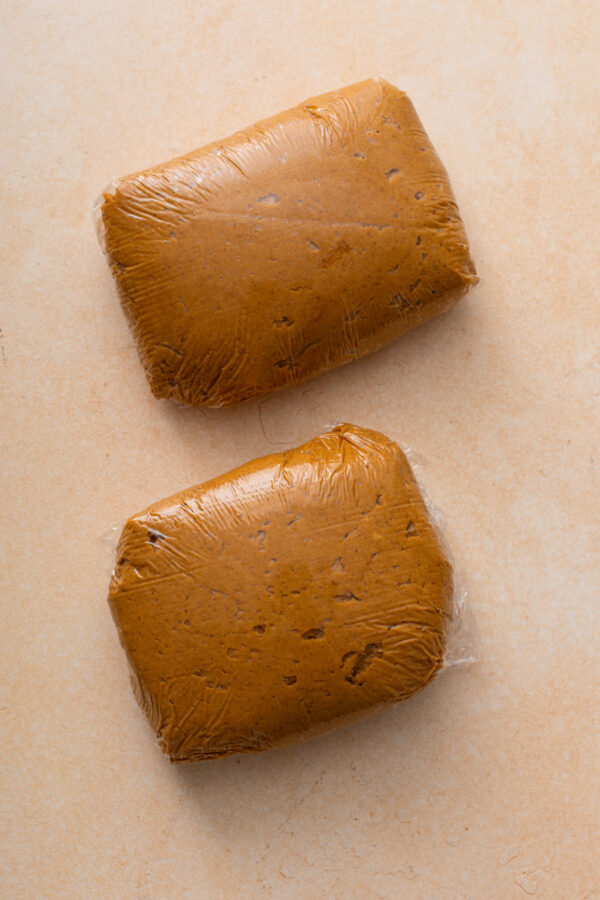 Dough for pumpkin molasses cookies split into two rectangles and wrapped in plastic wrap, ready to be chilled.