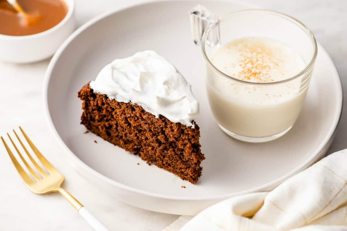 Slice of gingerbread cake topped with whipped cream next to a glass of eggnog