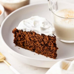 whipped-cream-topped gingerbread cake on a white plate next to a glass of eggnog