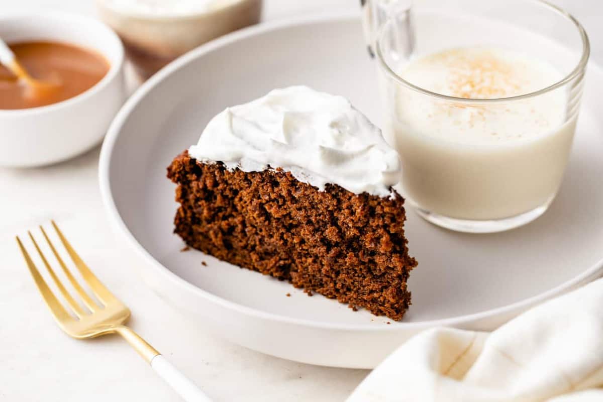 whipped-cream-topped gingerbread cake on a white plate next to a glass of eggnog