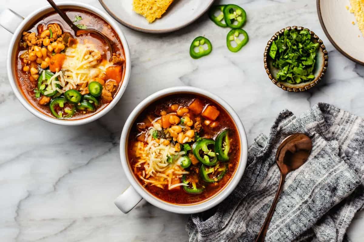 White bean chili in bowls on a countertop next to cornbread and sliced jalapenos