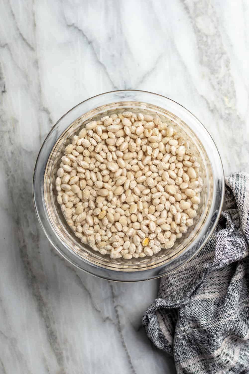 White beans soaking in a glass bowl on a marble countertop