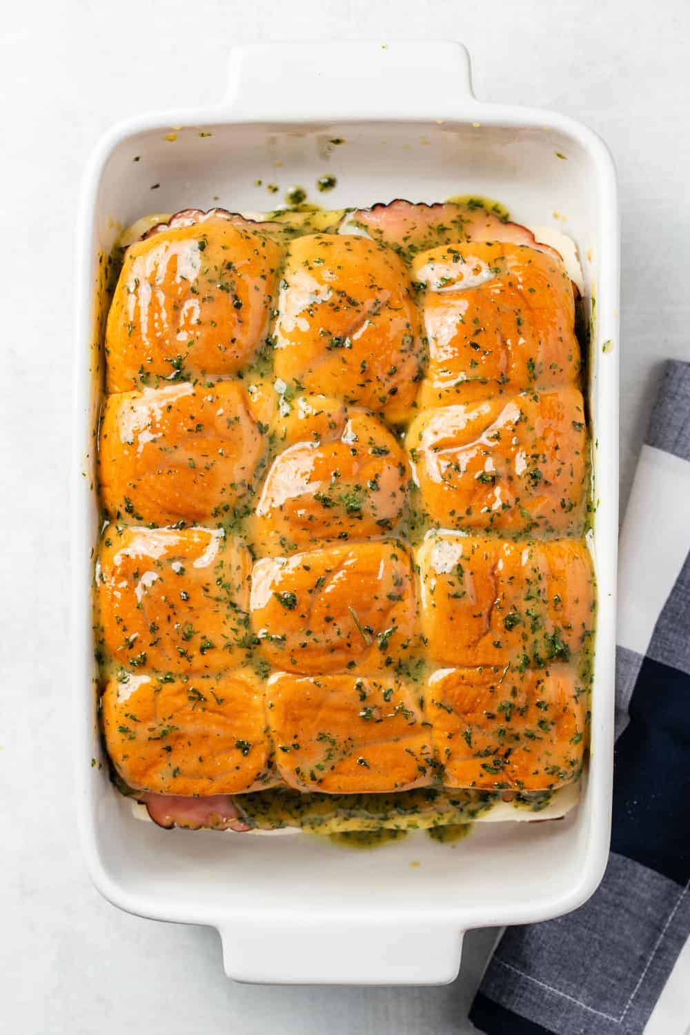 assembled ham and cheese sliders brushed with melted herb butter in a white baking dish, ready to be baked