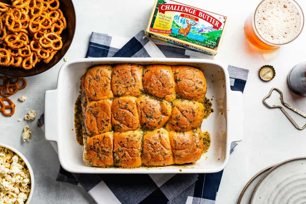 Baked ham and cheese sliders in a white baking dish surrounded by game day snacks