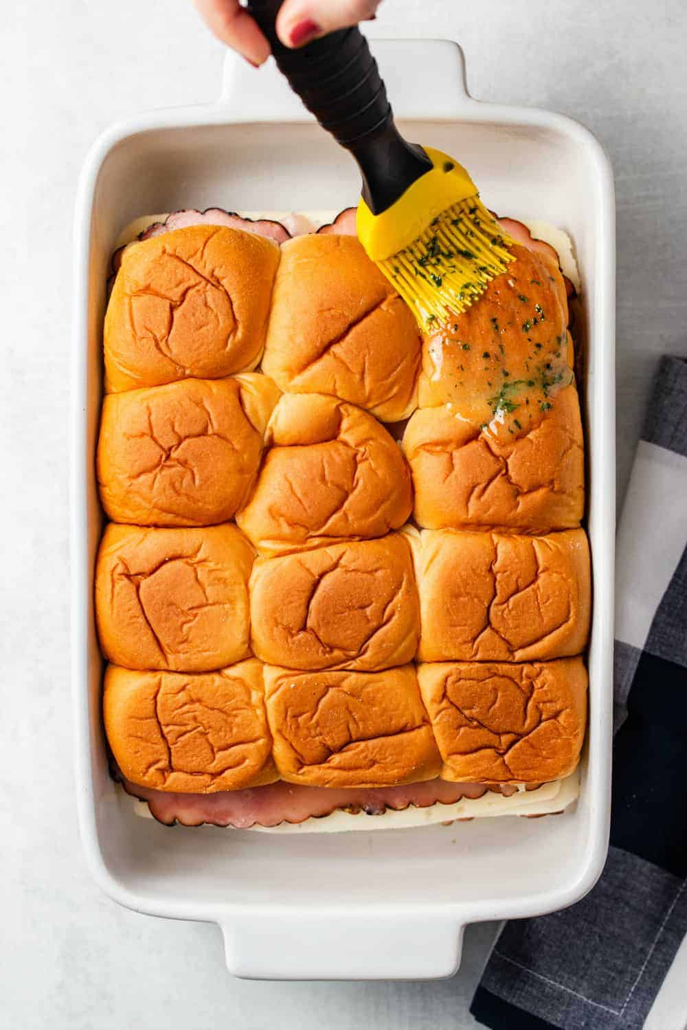 Brush adding melted herb butter to the top of hawaiian rolls for ham and cheese sliders in a white baking dish