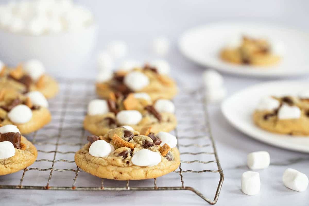 S'mores cookies lined up on a metal cooling rack with mini marshmallows scattered on the countertop