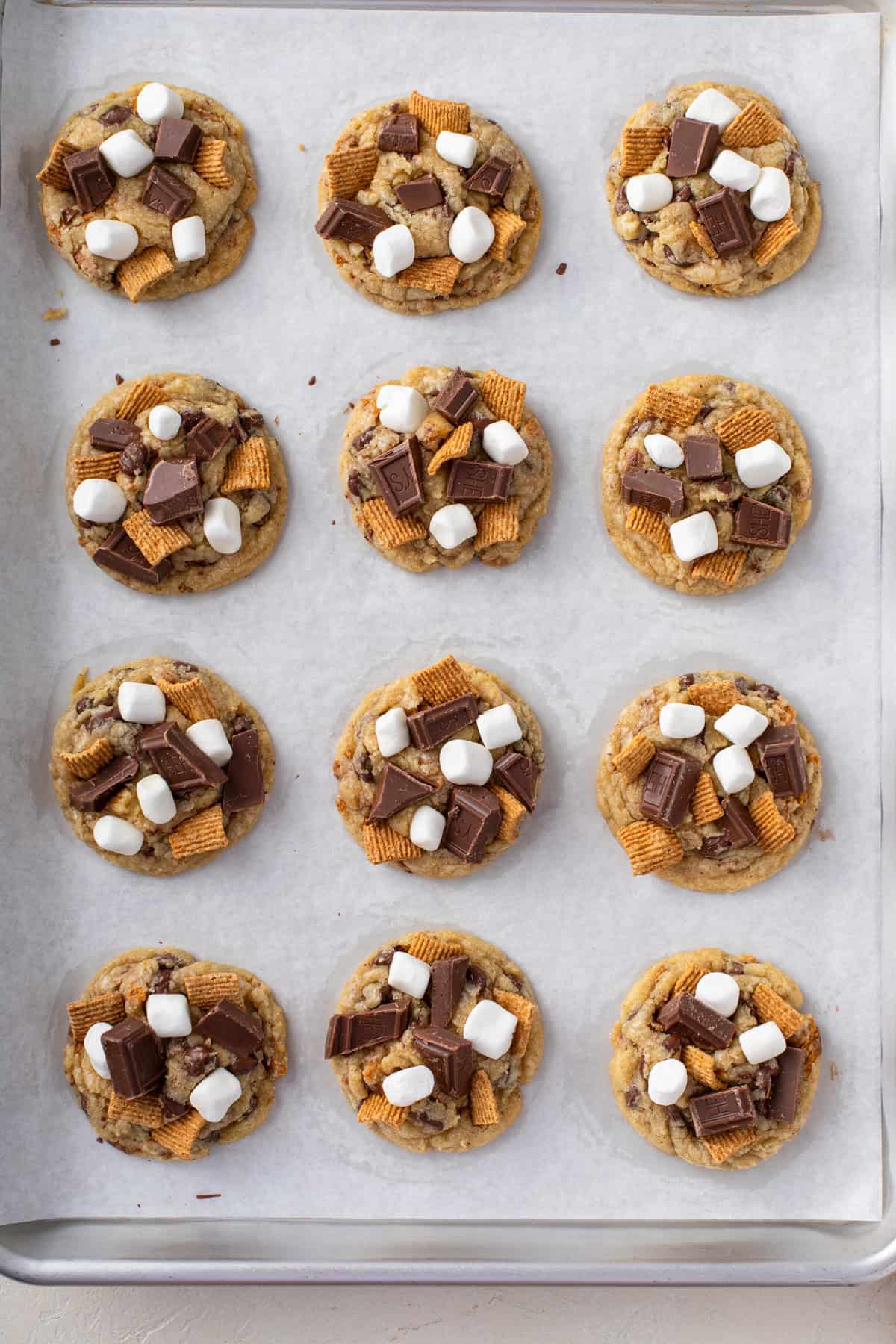 Partially baked s'mores cookies on a sheet tray topped with mini marshmallows, chocolate pieces, and graham cereal.