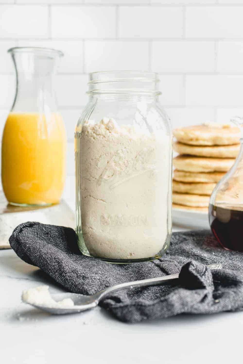 Glass jar of homemade Bisquick mix in front of a stack of Bisquick pancakes and a carafe of orange juice