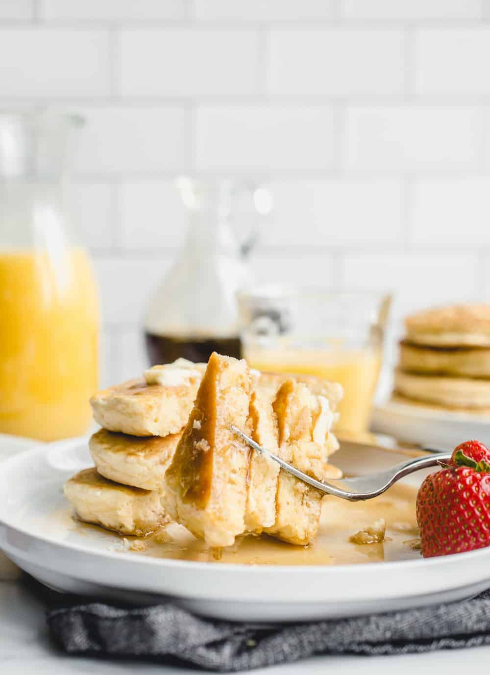 Fork taking a bite out of a stack of Bisquick pancakes on a white plate, with additional pancakes and orange juice in the background.